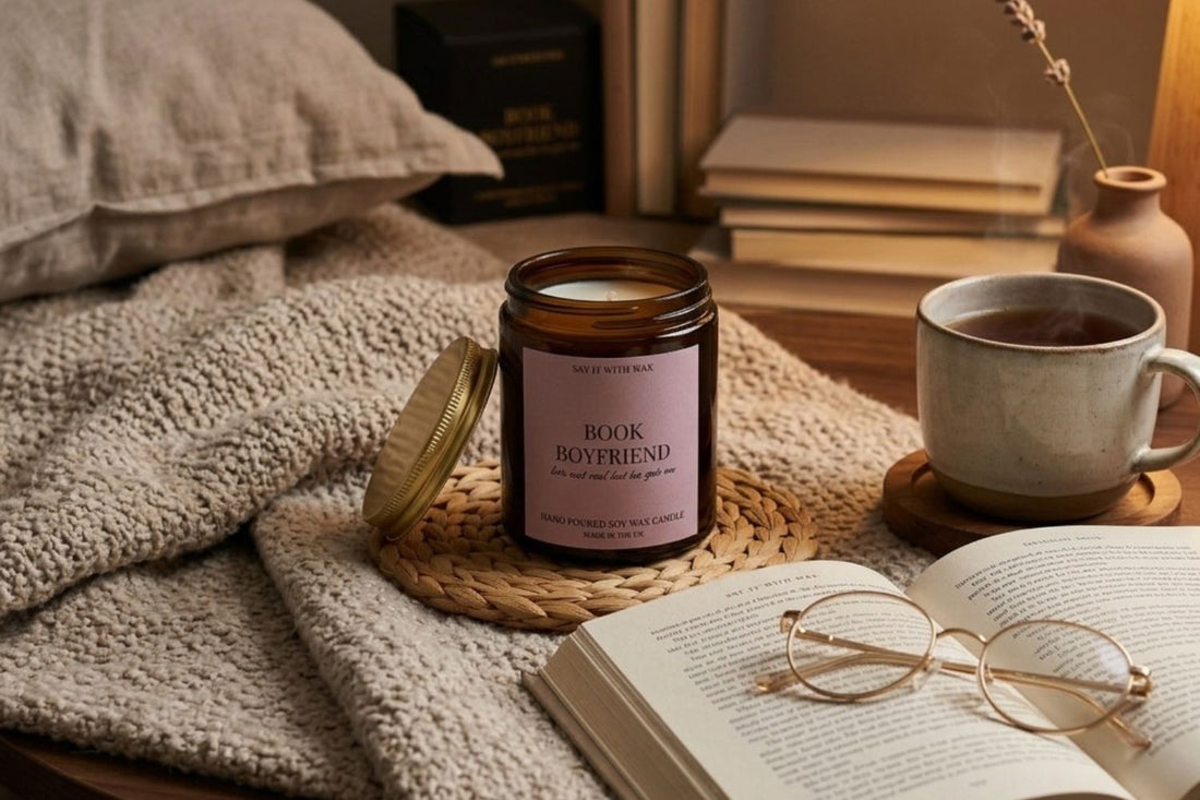 Book Boyfriend soy candle lit on a woven coaster beside an open book, glasses and a cup of tea, surrounded by knitted blankets in warm autumn light