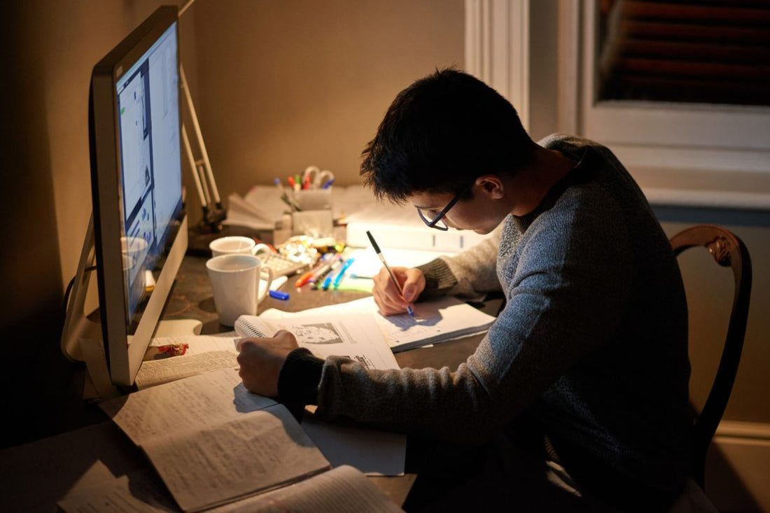 Student studying at a desk at night, writing in a notebook under warm lamp light, with a computer monitor, mugs and pens nearby