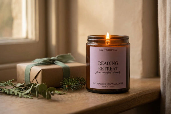 Lit Reading Retreat soy candle on a windowsill beside a plainly wrapped gift and sprigs of greenery, for the Budget Christmas Gifts Under £20 blog post.