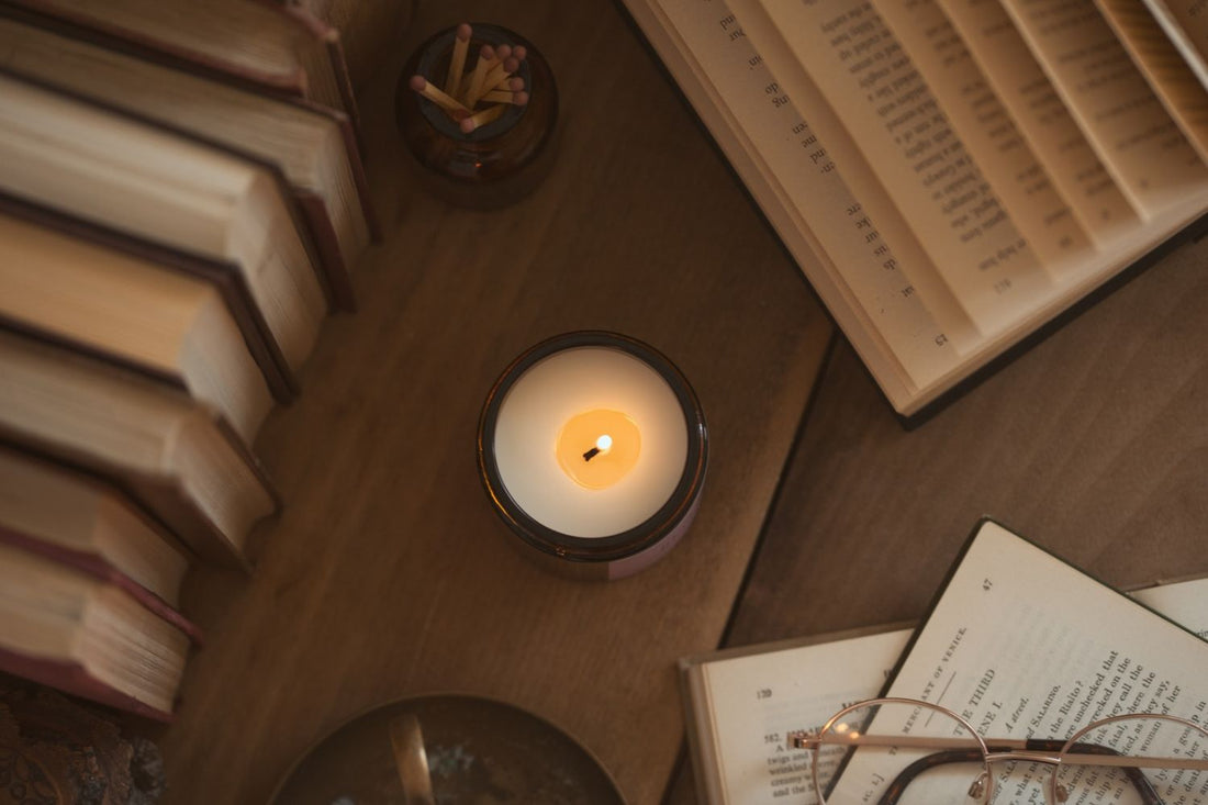 Overhead shot of a lit Say It With Wax soy candle in an amber jar on a wooden table, surrounded by open books, a jar of matches and reading glasses.