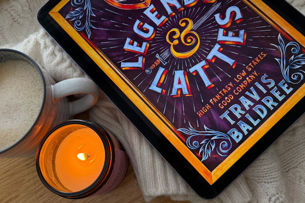 Lit soy candle beside a mug of coffee and a tablet showing the Legends & Lattes book cover, on a cream knitted blanket, for the Cosy Autumn Reading List blog post.