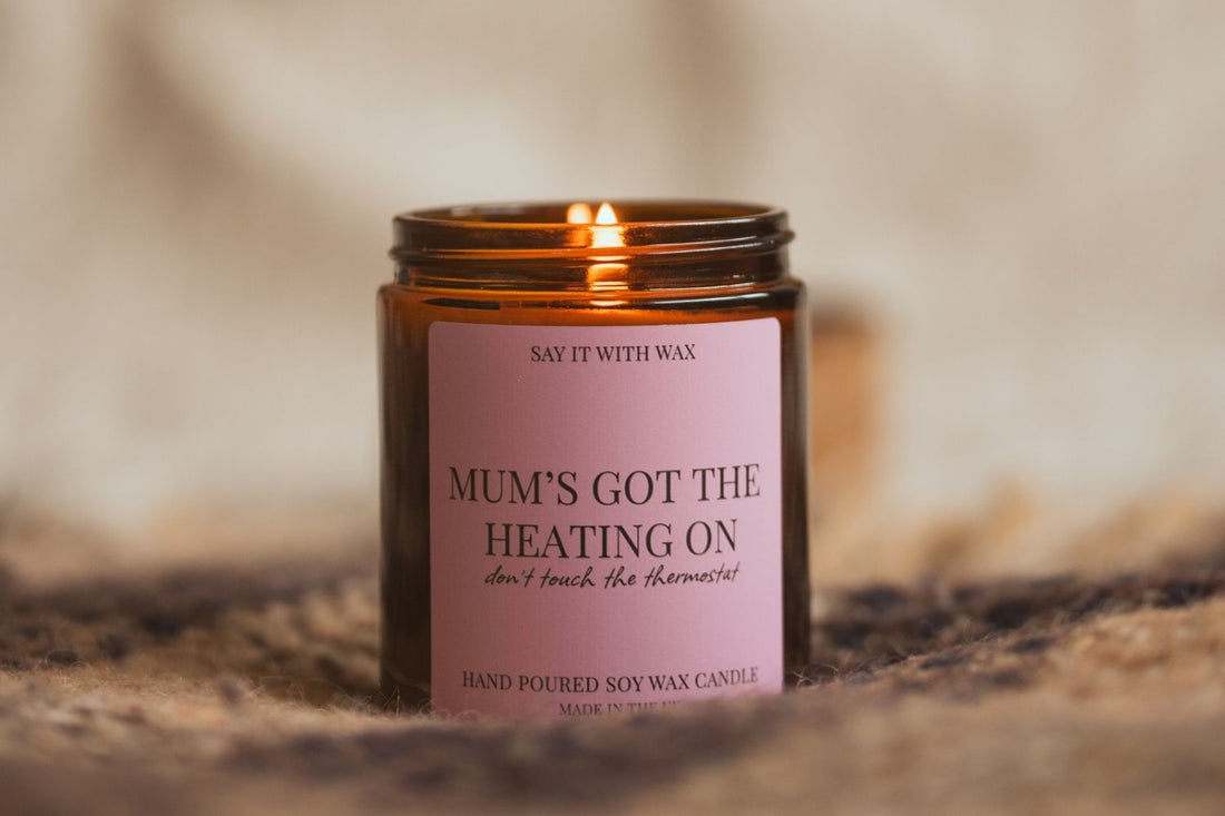 Lit Mum's Got the Heating On soy candle in an amber jar with a pink label, glowing on a knitted throw for the Empty Nest Gifts blog post.
