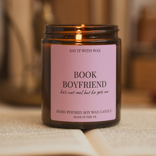 Say It With Wax Book Boyfriend soy candle burning, pink label reading "he's not real but he gets me", blurred books in the background.