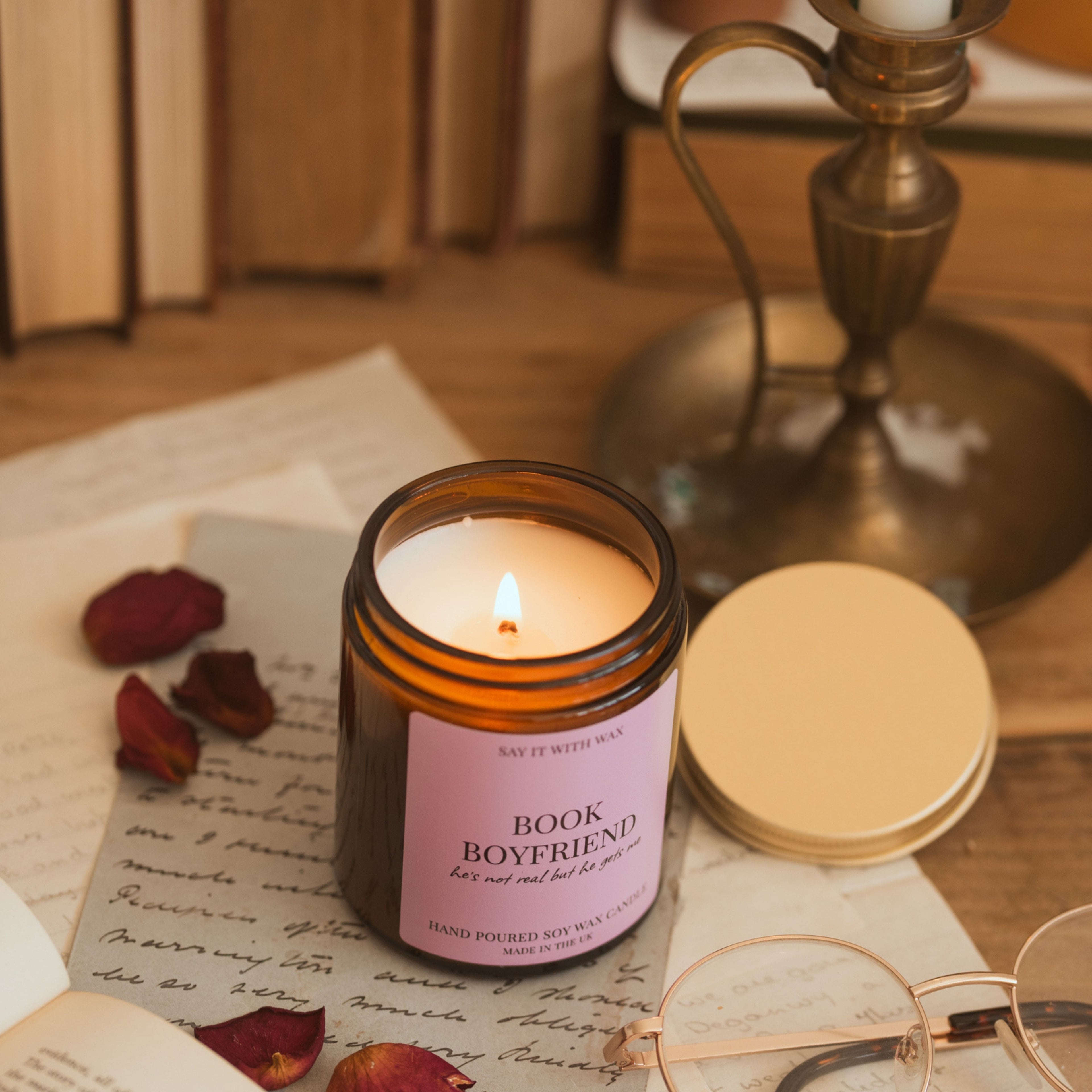 Lit Book Boyfriend candle surrounded by handwritten letters, dried rose petals and a brass ornament, with reading glasses in the foreground.