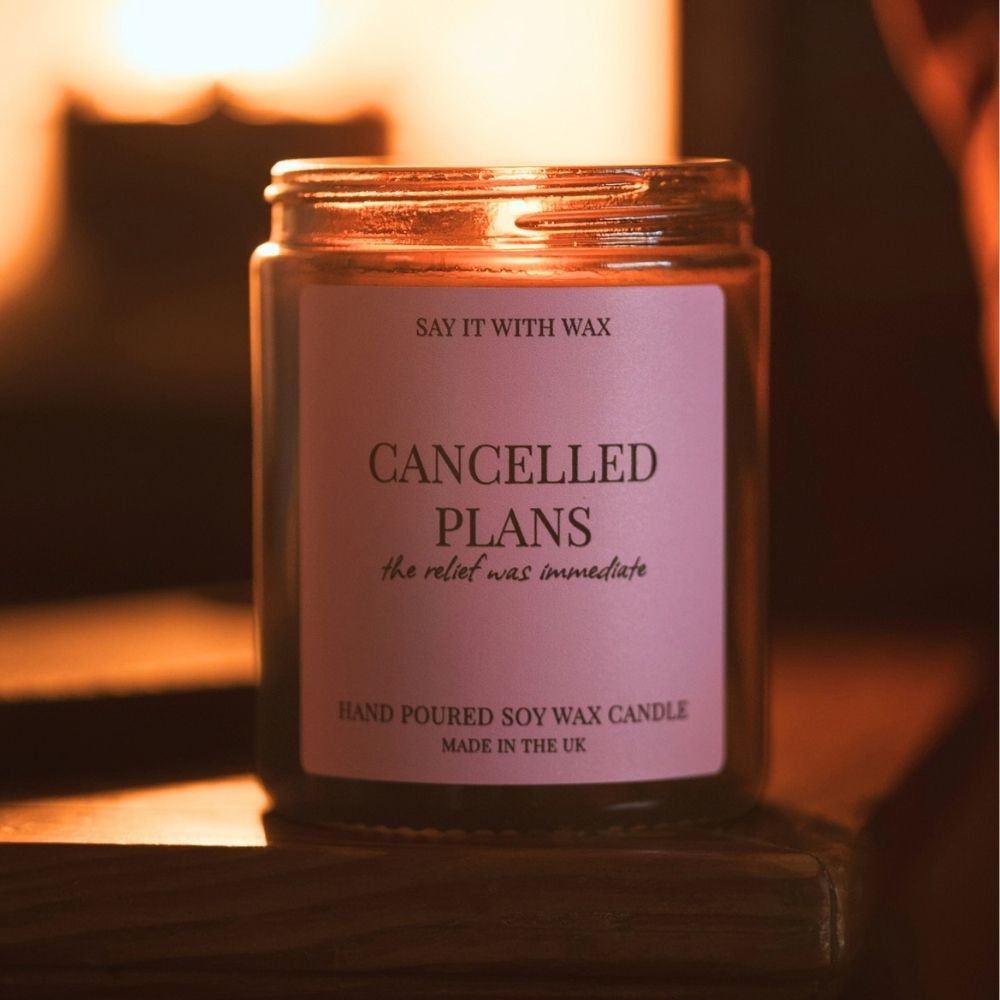 Lit Cancelled Plans hand poured soy wax candle in an amber glass jar with a pink label reading "the relief was immediate", glowing on a wooden shelf