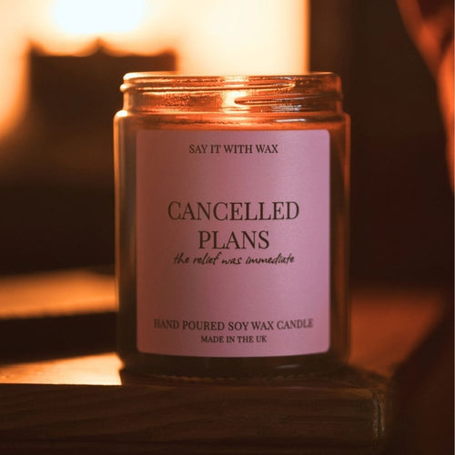 Lit Cancelled Plans hand poured soy wax candle in an amber glass jar with a pink label reading "the relief was immediate", glowing on a wooden shelf