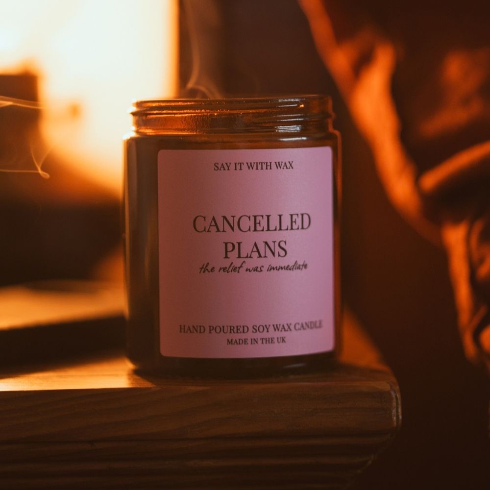 Cancelled Plans soy wax candle in an amber jar with a pink label, just blown out with a trail of smoke rising in a warm, cosy room
