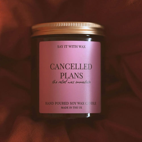Cancelled Plans soy wax candle with a gold lid and pink label, resting on soft rust-coloured fabric