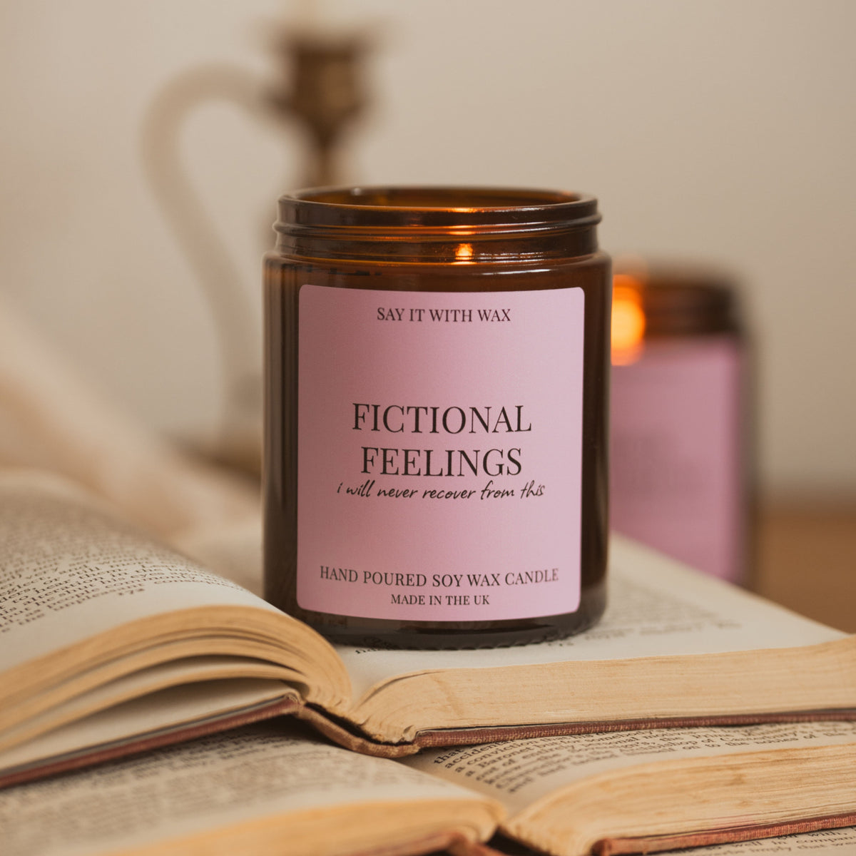An amber glass soy wax candle jar with a pink label reading 'FICTIONAL FEELINGS' sits on an open book, with a softly blurred candle and books in the background.