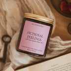 A hand-poured soy wax candle in a dark glass jar with a gold metal lid and a dusty pink label reading 'FICTIONAL FEELINGS'. The candle is styled on a bed of cream-colored fabric with an antique key, an open book, and a small wooden dish in the background.