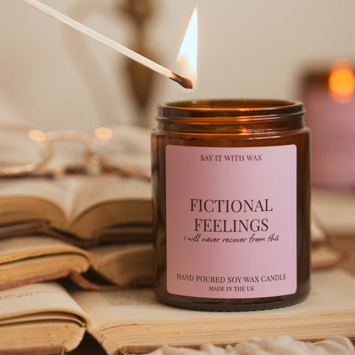 An amber glass soy wax candle jar with a pink label reading 'FICTIONAL FEELINGS' and a lit match hovering above it, styled on open books with a soft warm background.