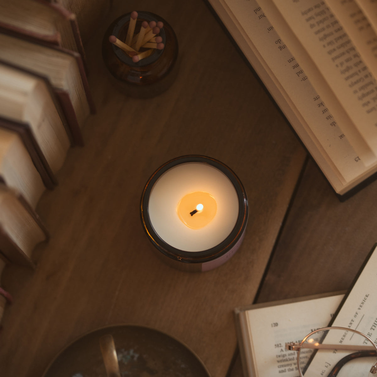 Top-down view of a lit candle on a wooden surface, with a jar of matches, an open book and glasses nearby.