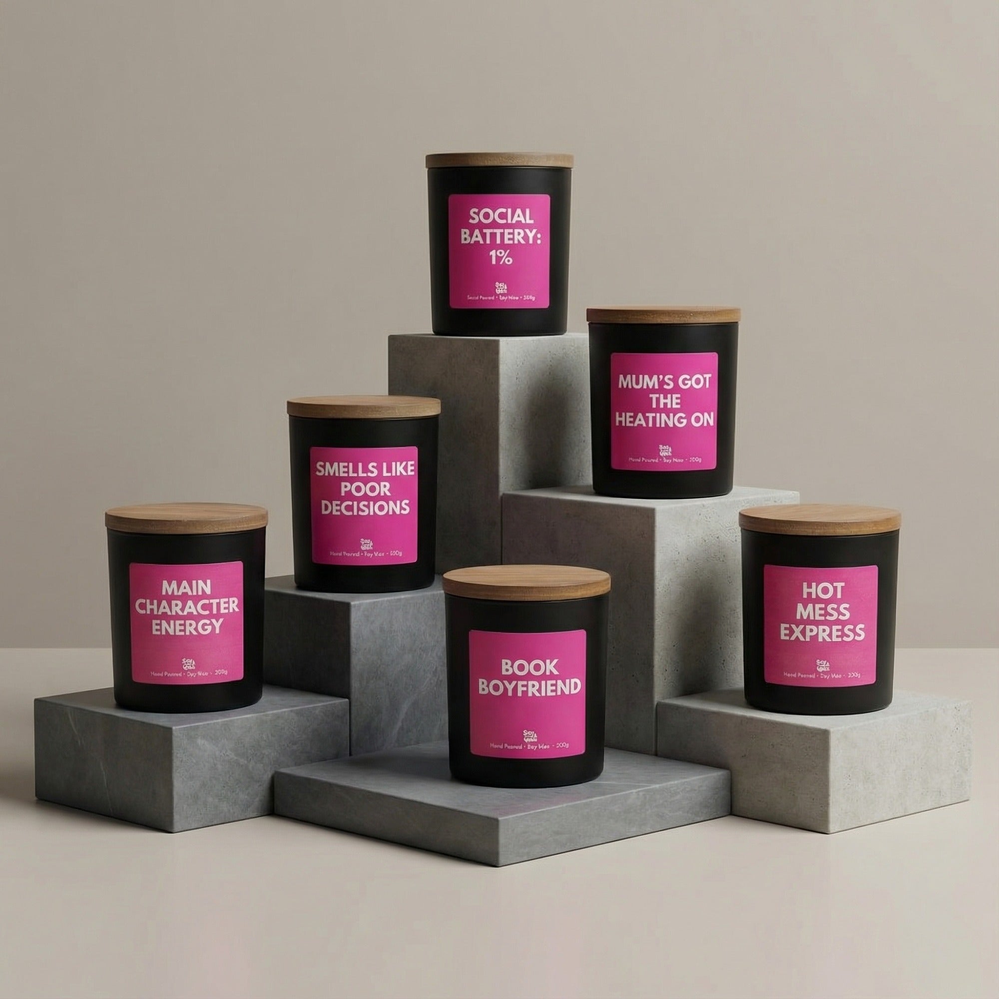 Six black candles with wooden lids and bright pink labels are displayed on staggered grey stone blocks against a soft beige background. The candles are arranged in a modern, sculptural layout. The labels read: “Social Battery: 1%,” “Mum’s Got the Heating On,” “Hot Mess Express,” “Book Boyfriend,” “Smells Like Poor Decisions,” and “Main Character Energy.” The overall look is clean, minimal and stylish, showcasing the collection in a bold, cohesive way.