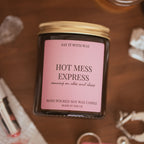 Hot Mess Express soy candle with gold lid on, styled on a wooden surface beside lipstick, jewellery and matches.