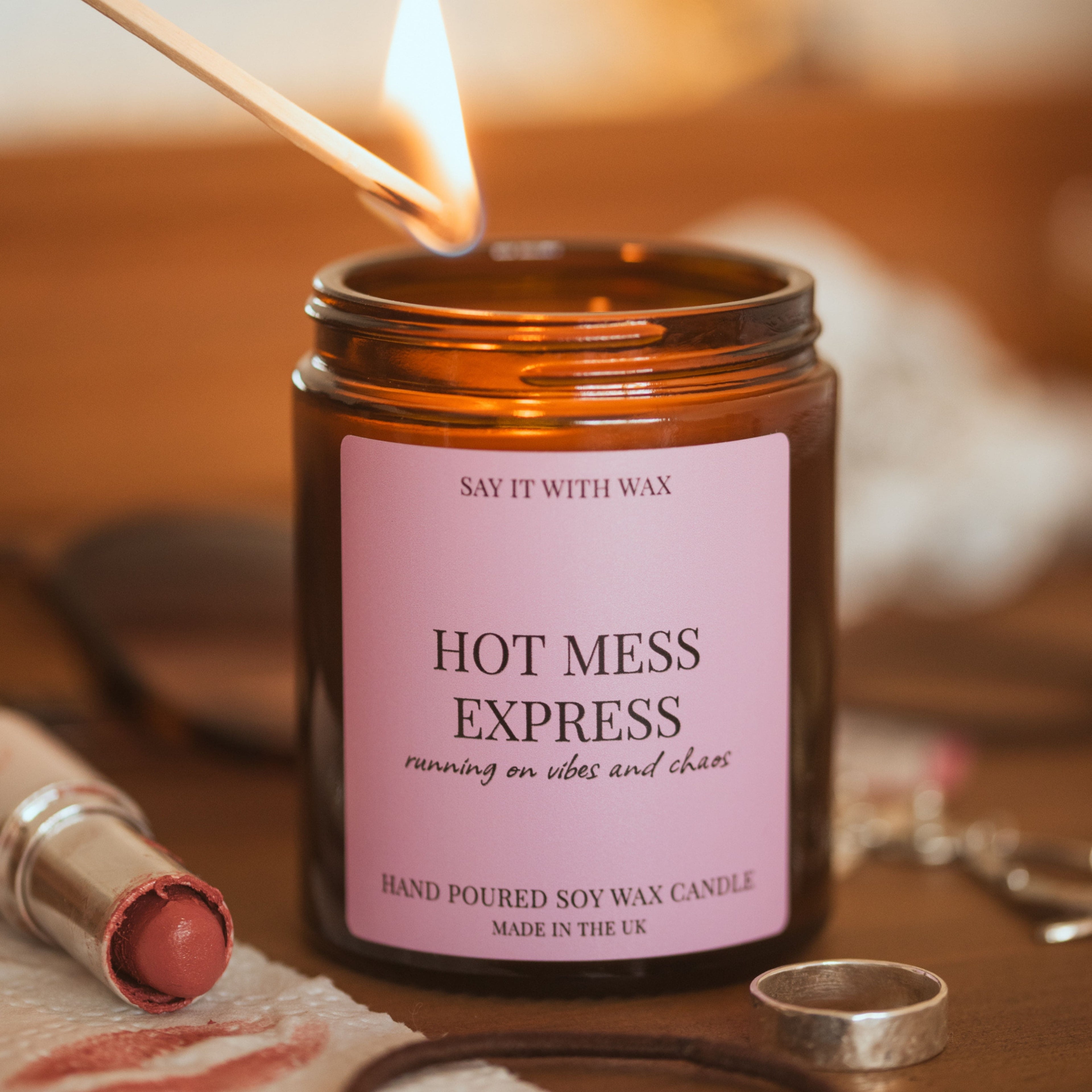 A lit match held above the open Hot Mess Express candle by Say It With Wax, with a lipstick, rings and a soft blanket blurred in the background.