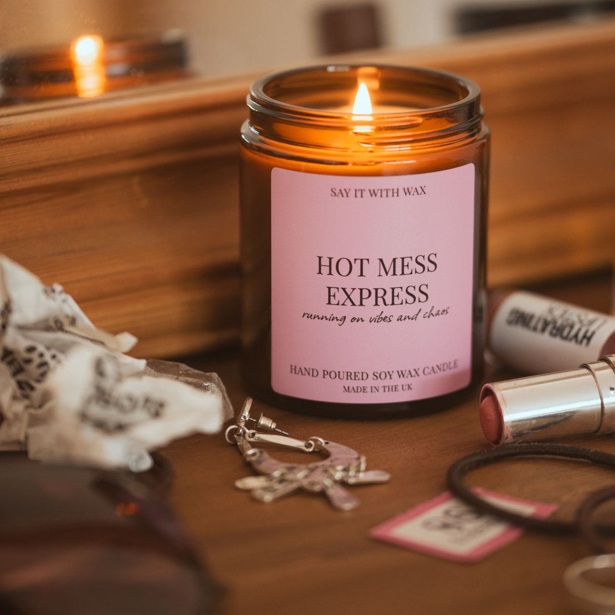 Lit Hot Mess Express candle surrounded by lipstick, hair ties, keys and a second candle glowing in the background, styled like a lived-in dressing table.