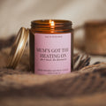 Lit Mum's Got the Heating On candle by Say It With Wax with its gold lid propped against the jar, nestled in a woven throw.