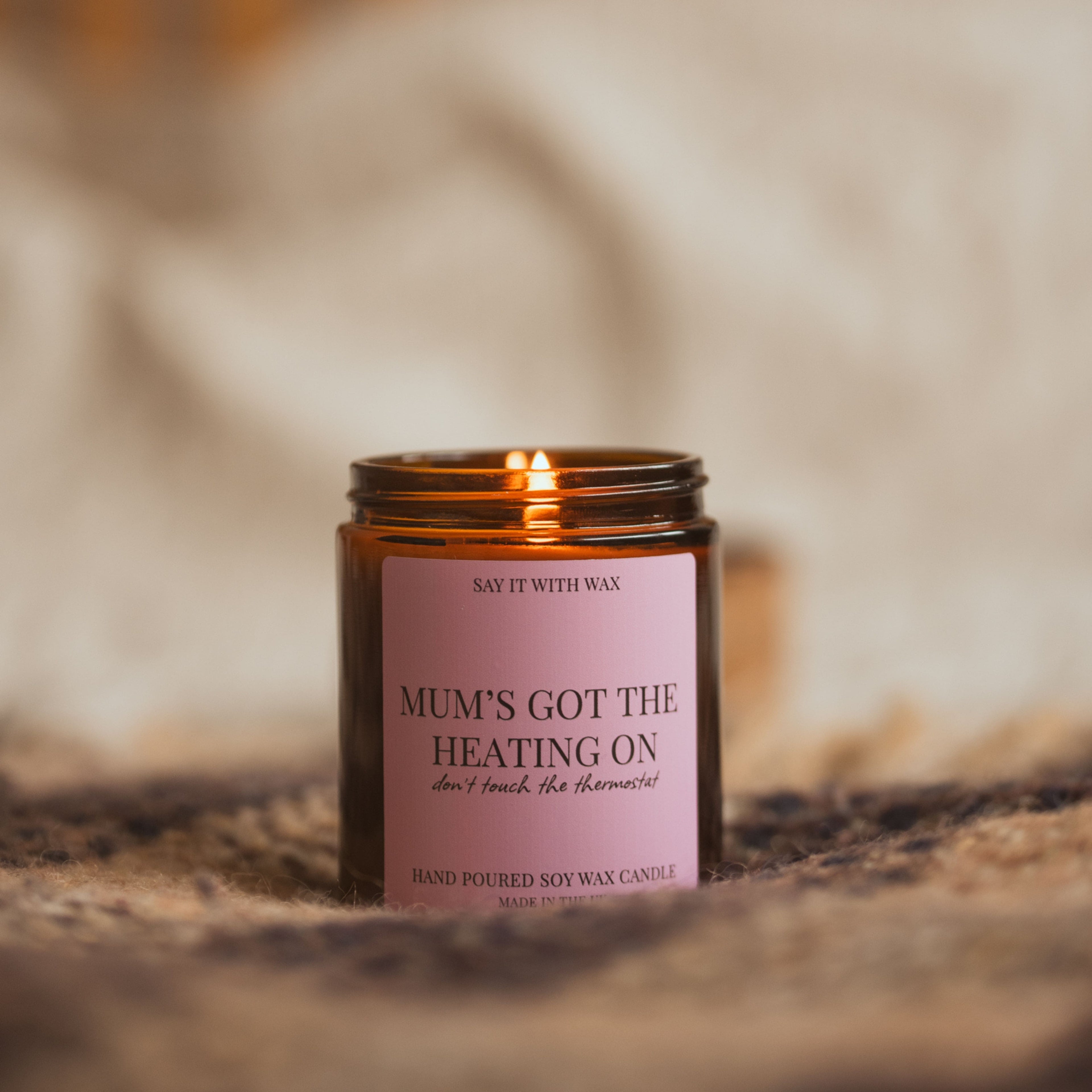 Lit Mum's Got the Heating On candle glowing against a warm, blurred cosy background, styled on a woven throw.
