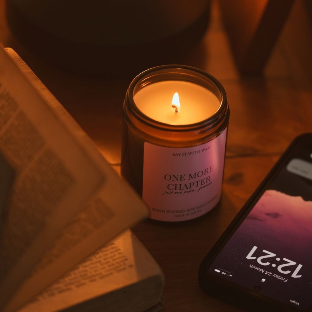 One More Chapter hand poured soy wax candle by Say It With Wax, lit and glowing beside an open book and a phone showing 12:21 on a wooden table
