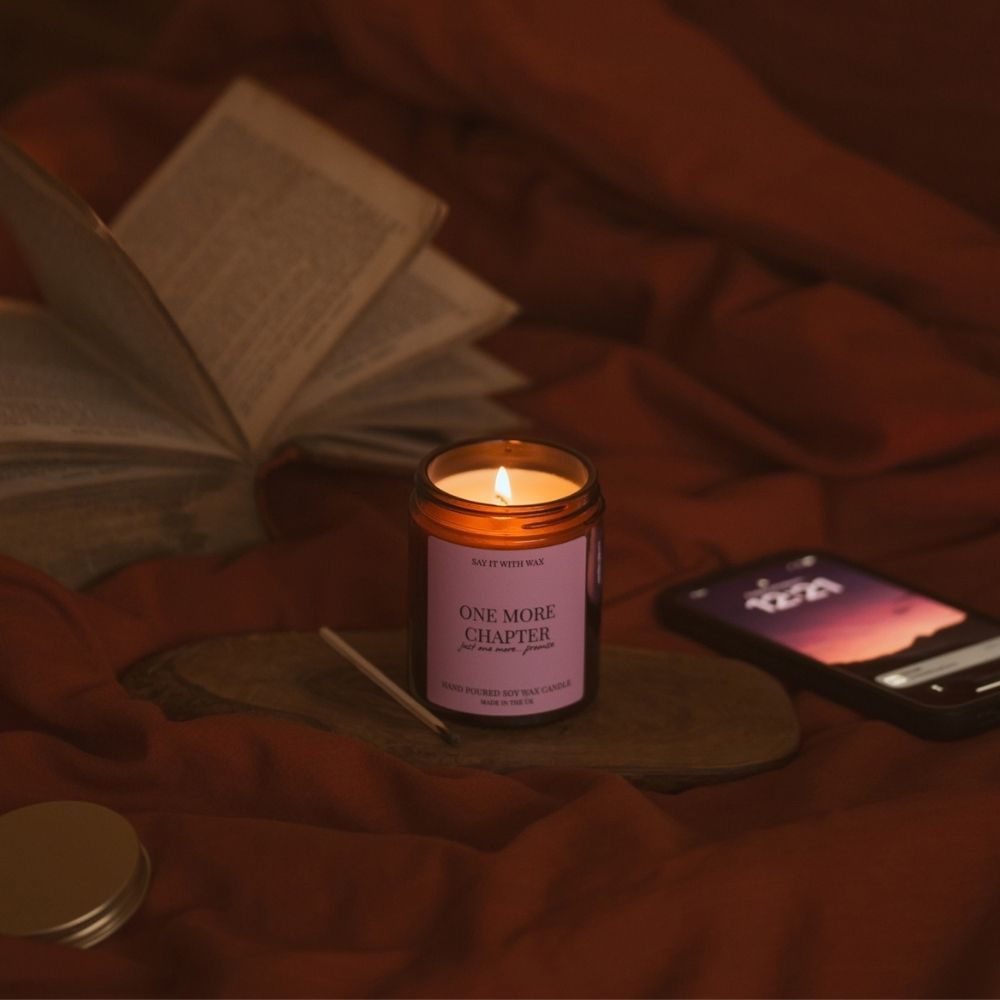 One More Chapter soy wax candle burning on a wooden board among cosy red bedding, with an open book, a phone and a spent match beside it