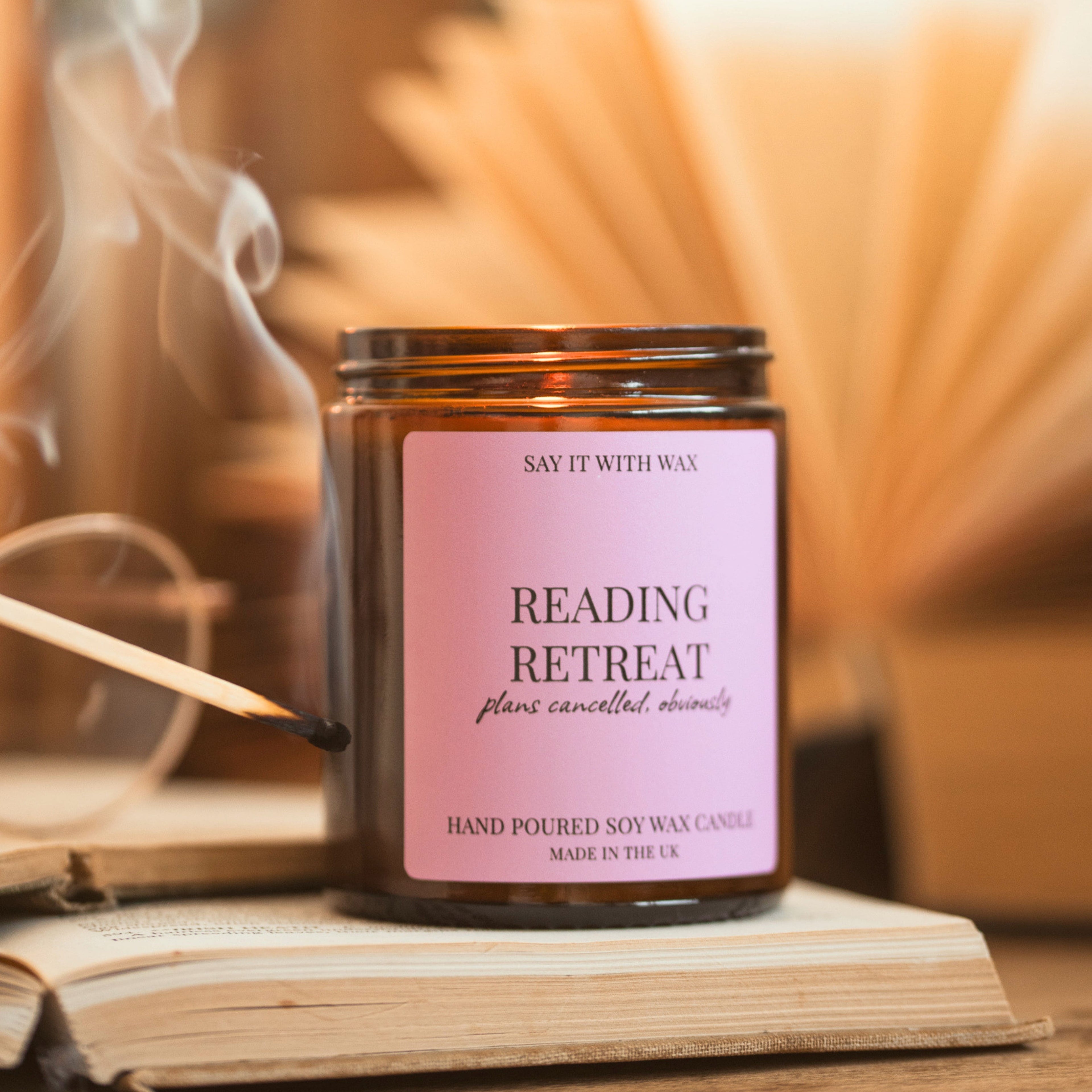 A just-blown-out match beside the lit Reading Retreat candle by Say It With Wax, resting on an open book with glasses and fanned book pages behind it.