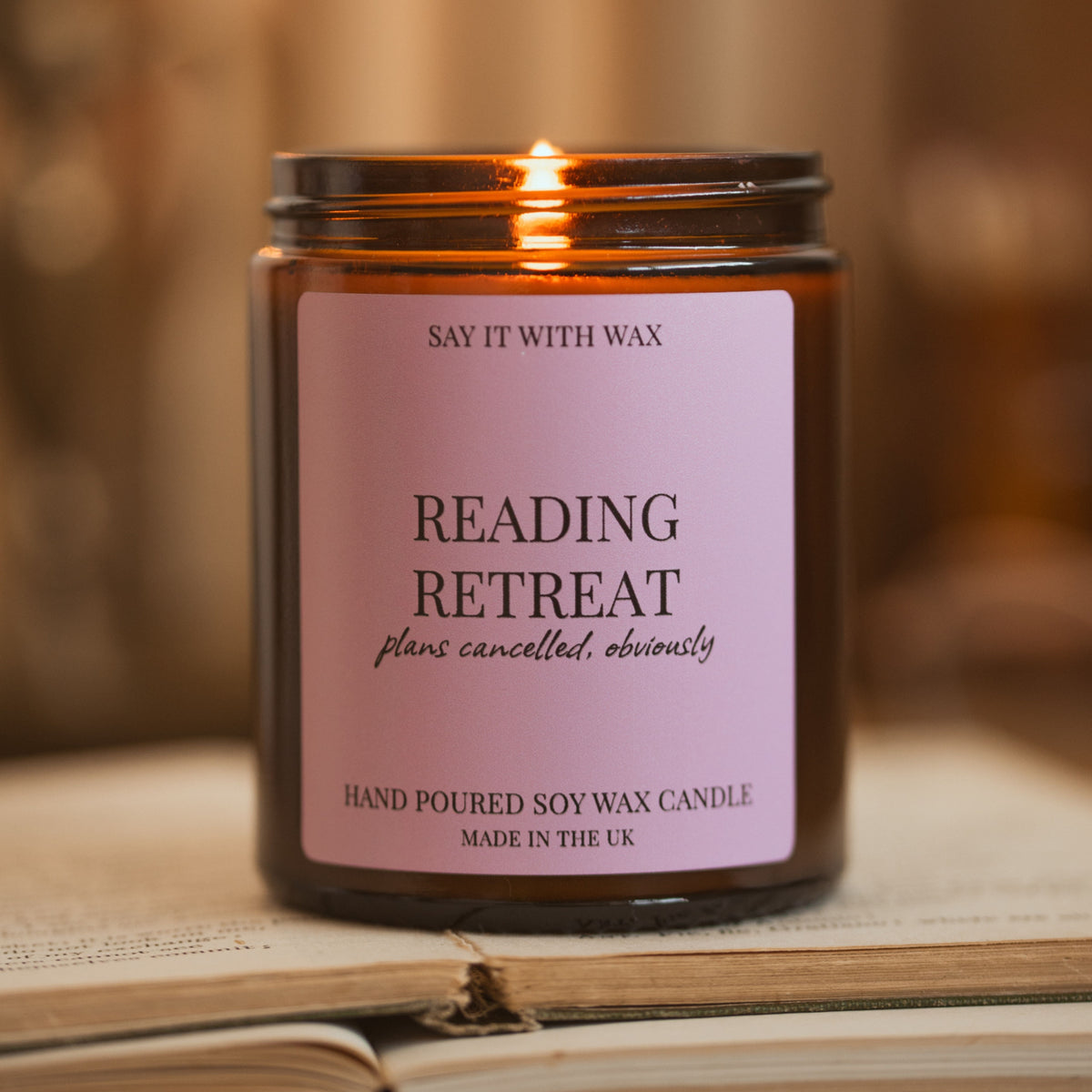 Say It With Wax Reading Retreat candle burning, pink label reading "plans cancelled, obviously", resting on an open book with a soft warm background.