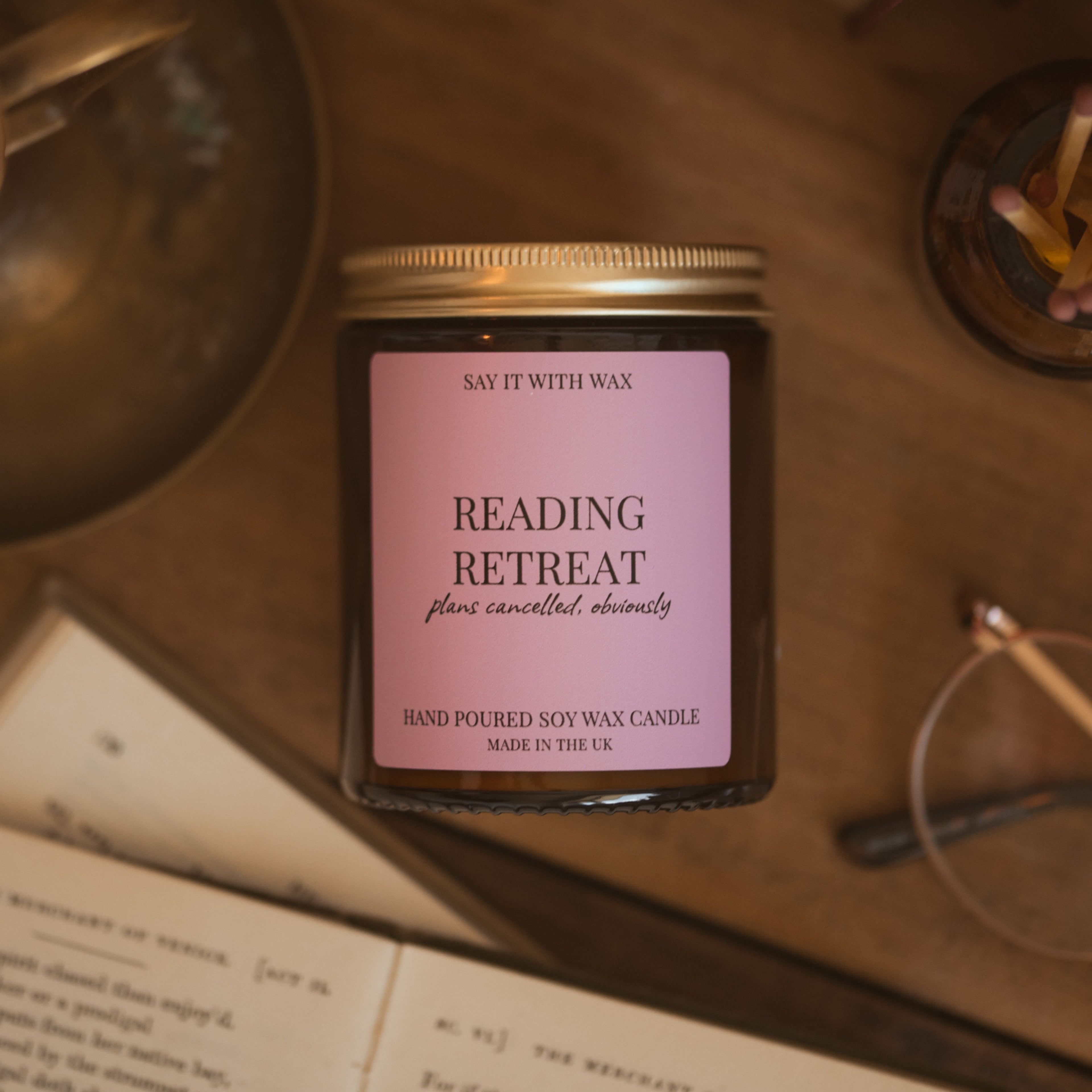 Reading Retreat candle with gold lid on, styled on a wooden desk beside handwritten pages, glasses and a jar of matches.