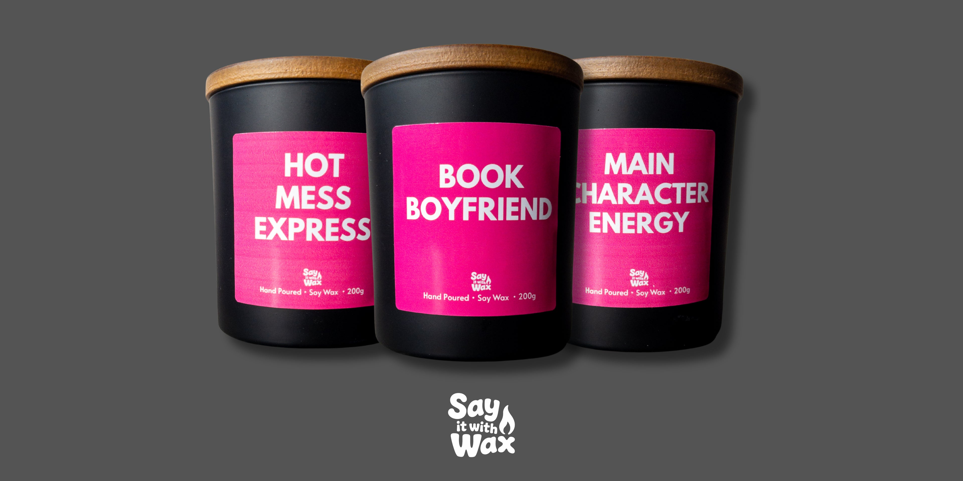Three black candles with pink labels on a gray background, featuring the brand 'Say Your Wax'.