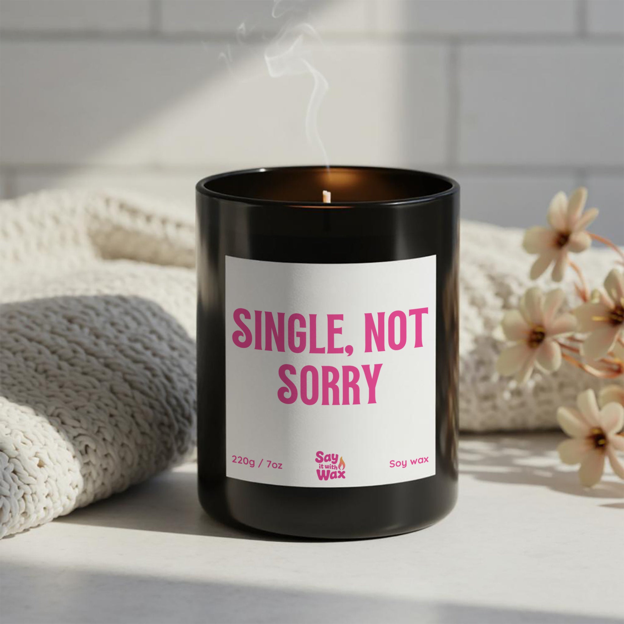 Single, Not Sorry” soy candle in black glass vessel with pink label, hand-poured by Say it With Wax, styled with flowers on white surface.