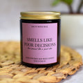 Smells Like Poor Decisions soy wax candle with a pink label on a woven mat