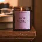 Lit Social Battery: 1% candle on a rustic wooden ledge, soft warm background with blurred glasses and books.
