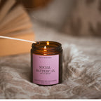 A lit match held above the open Social Battery: 1% candle, smoke curling upward, styled on soft fabric with dried palm leaves in the background.