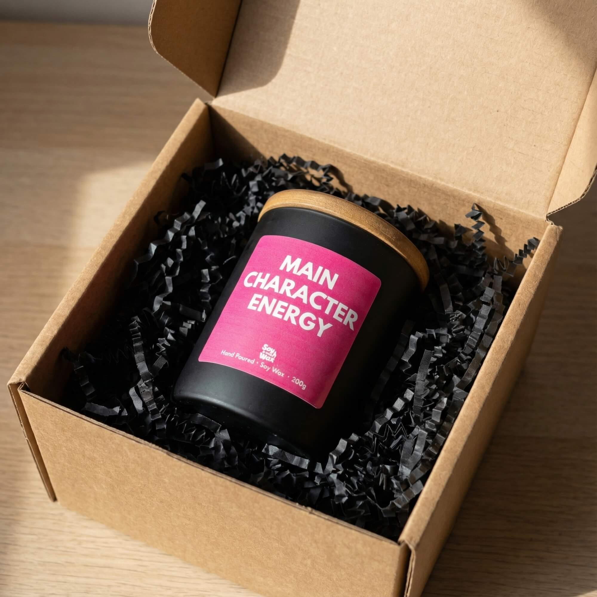 A black candle with a wooden lid and a bright pink label reading “Main Character Energy” is displayed inside an open kraft gift box filled with black shredded paper. The box sits on a light surface alongside brown wrapping tissue, a handwritten “Thank You” card, and a pair of vintage-style scissors. Soft natural light creates gentle shadows, giving the scene a cosy, premium unboxing feel.