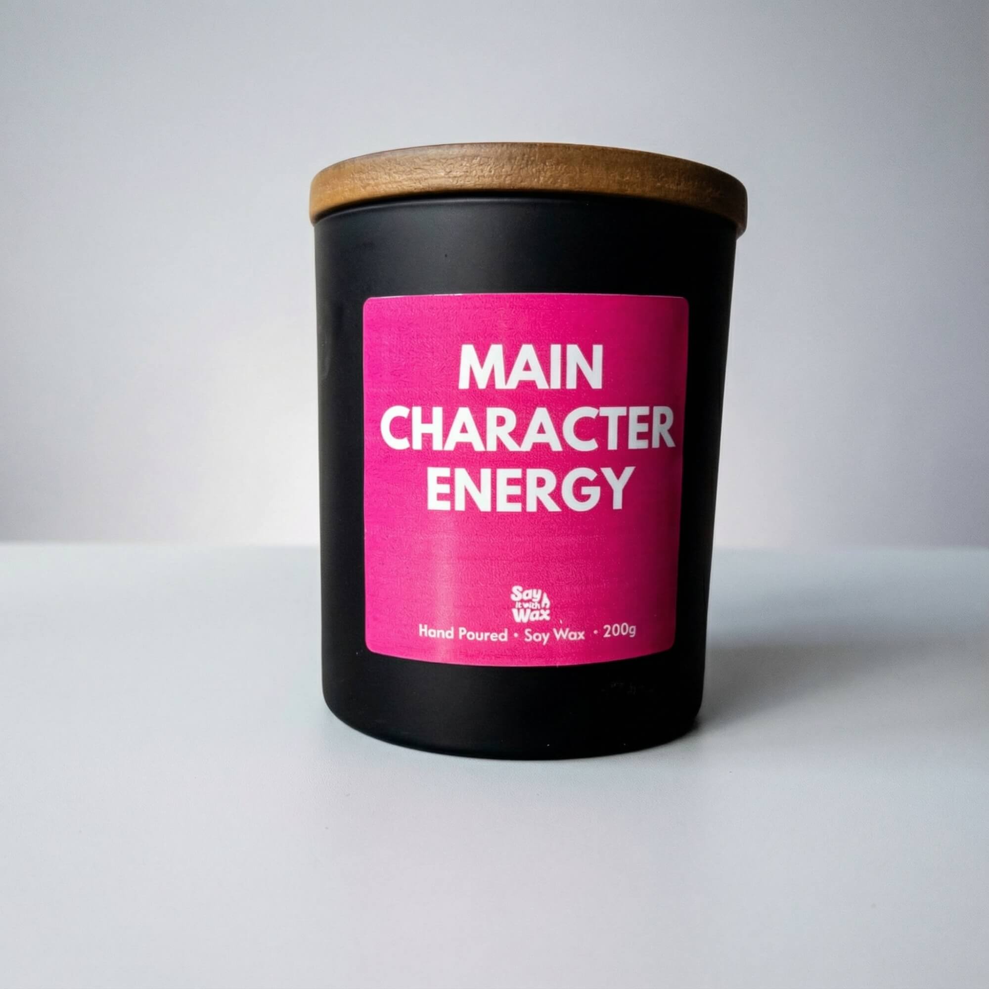 A black candle with a wooden lid and a bright pink label reading “Main Character Energy” is photographed against a simple light grey background. The candle is centred in the frame on a smooth white surface, with soft lighting creating gentle shadows for a clean, minimal product shot.