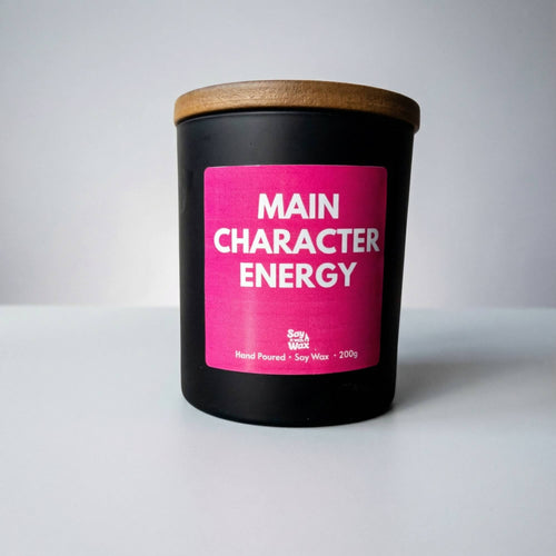 A black candle with a wooden lid and a bright pink label reading “Main Character Energy” is photographed against a simple light grey background. The candle is centred in the frame on a smooth white surface, with soft lighting creating gentle shadows for a clean, minimal product shot.