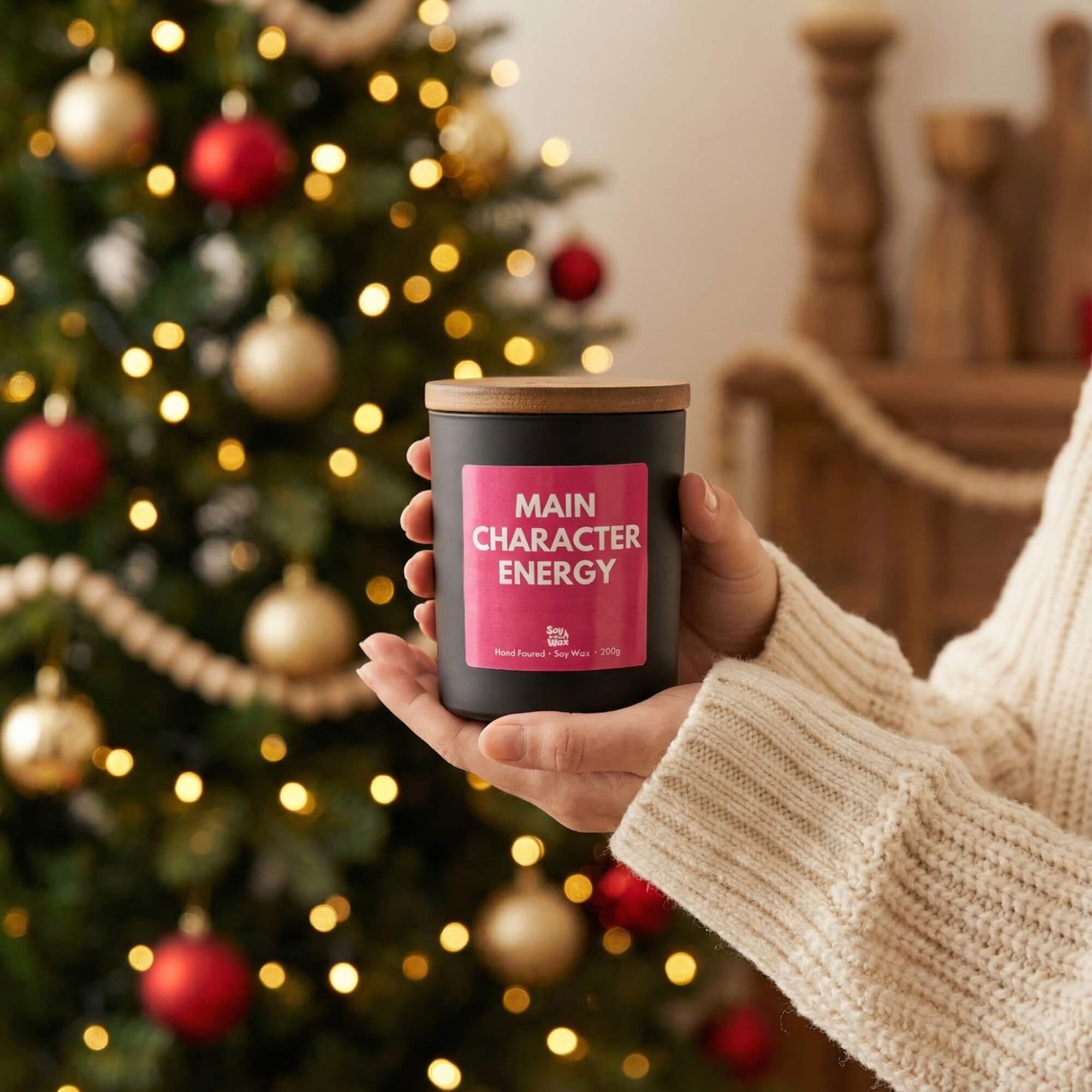 A person wearing a chunky cream knit jumper holds a black candle with a wooden lid and a bright pink label reading “Main Character Energy.” Behind them, a decorated Christmas tree glows with soft golden lights and red and gold baubles, creating a festive, cosy background.