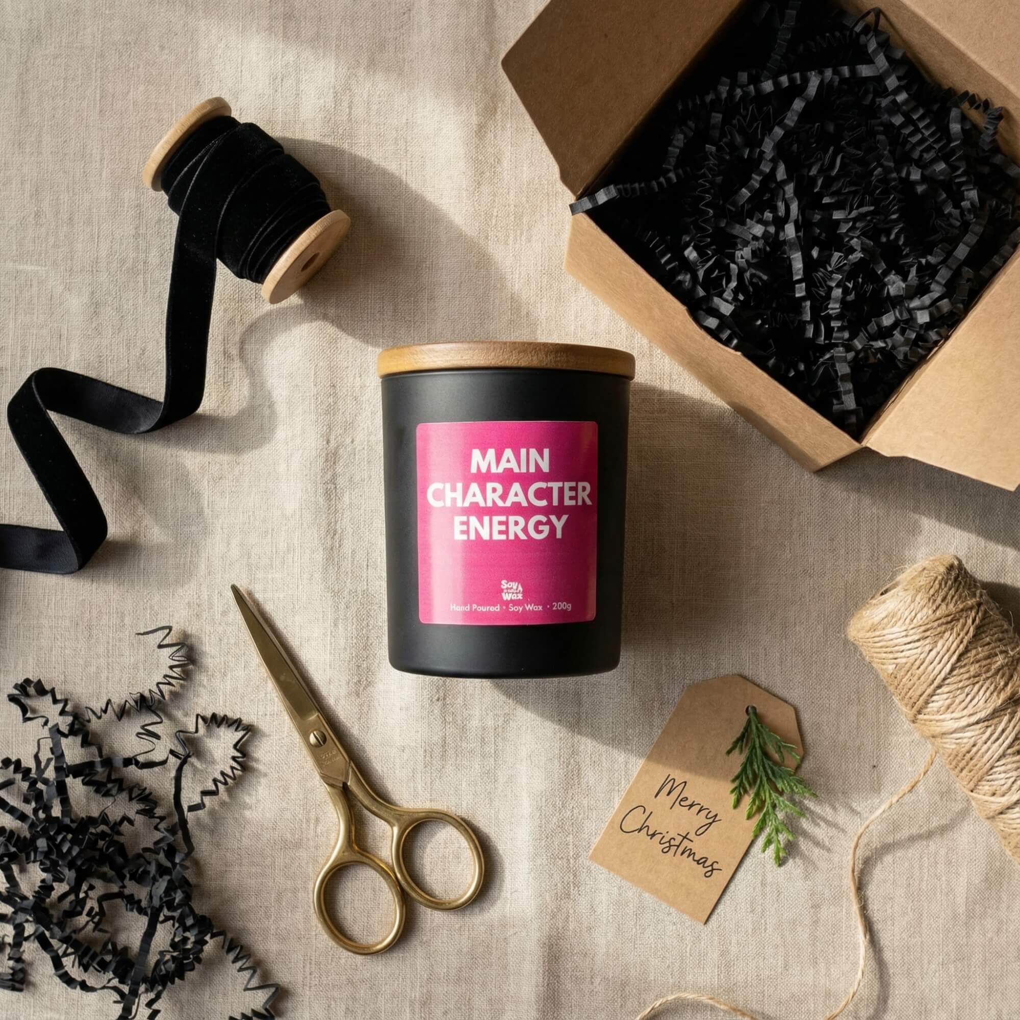 A black candle with a wooden lid and a bright pink label reading “Main Character Energy” is placed on a neutral fabric surface surrounded by Christmas gift-wrapping items. Nearby are gold scissors, black ribbon, a spool of twine, black shredded paper, an open kraft gift box, and a kraft gift tag that reads “Merry Christmas” with a small green sprig attached. Soft natural light creates warm shadows, giving the scene a cosy, festive feel.