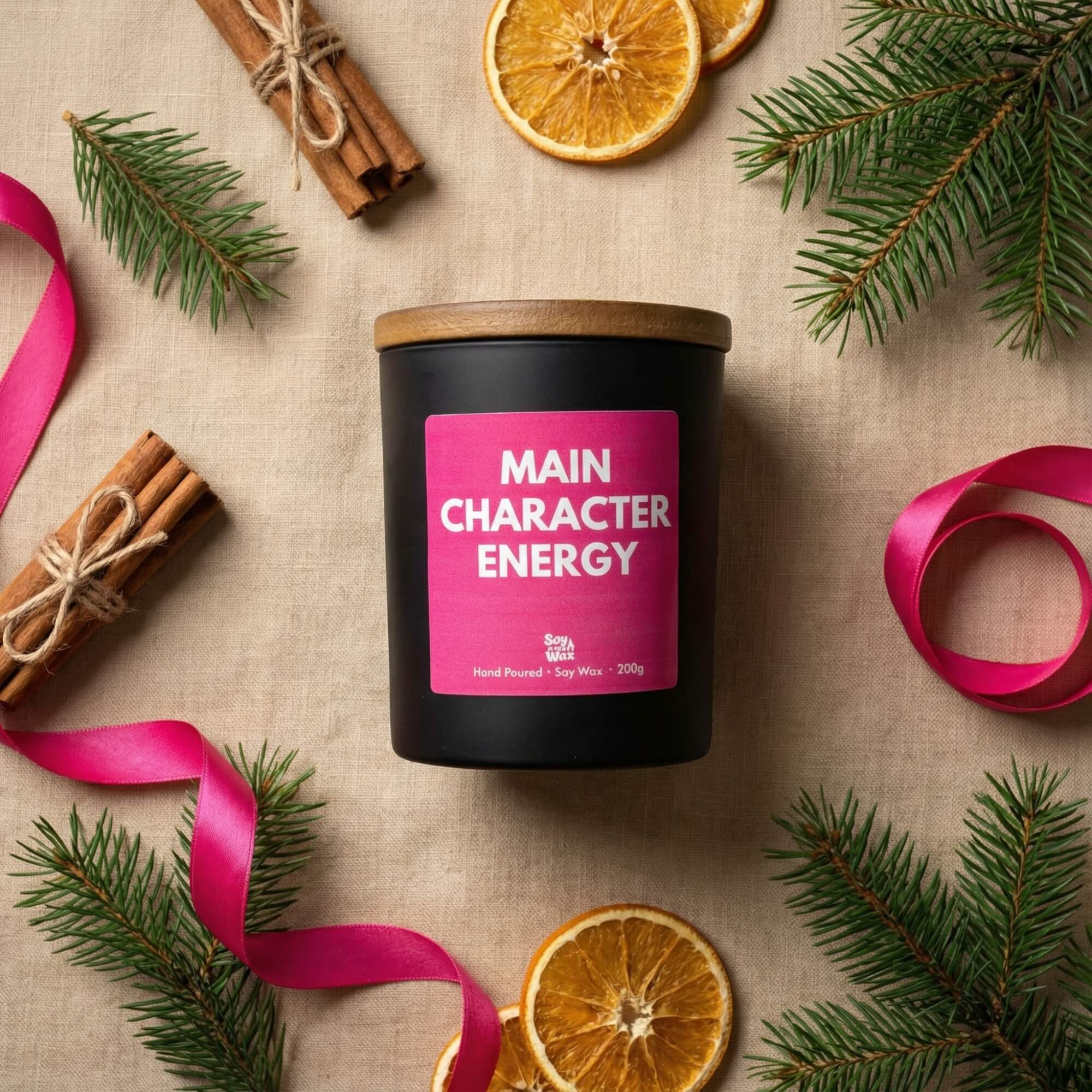 A black candle with a wooden lid and a bright pink label reading “Main Character Energy” is placed on a neutral fabric surface. Surrounding it are Christmas-themed props: sprigs of evergreen, dried orange slices, cinnamon sticks tied with twine, and a length of vibrant pink ribbon. The scene feels warm, seasonal, and gift-ready.