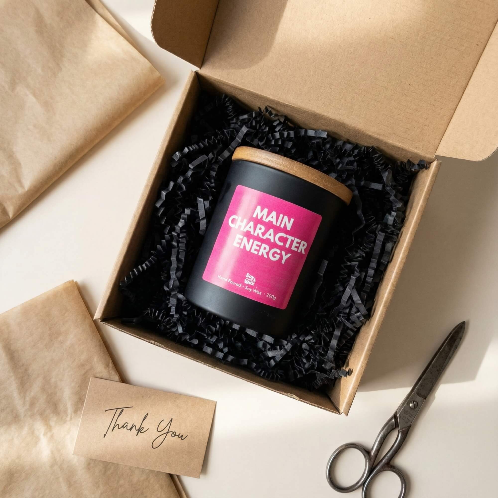 A black candle with a wooden lid and a bright pink label reading “Main Character Energy” is displayed inside an open kraft gift box filled with black shredded paper. The box sits on a light surface alongside brown wrapping tissue, a handwritten “Thank You” card, and a pair of vintage-style scissors. Soft natural light creates gentle shadows, giving the scene a cosy, premium unboxing feel.