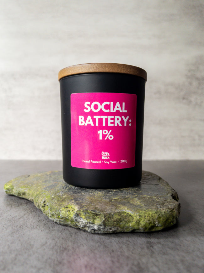 Candle with a pink label reading 'Social Battery: 1%' on a stone surface.